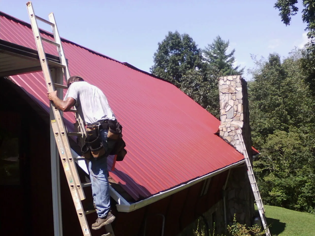Roofer on a ladder beside a red metal roof and stone chimney — Soffit & Fascia Repair by Roofing Brothers in Lander